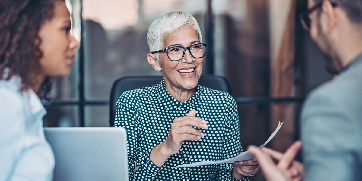 Woman with short white hair and glasses speaks to a man and woman while holding a paper document