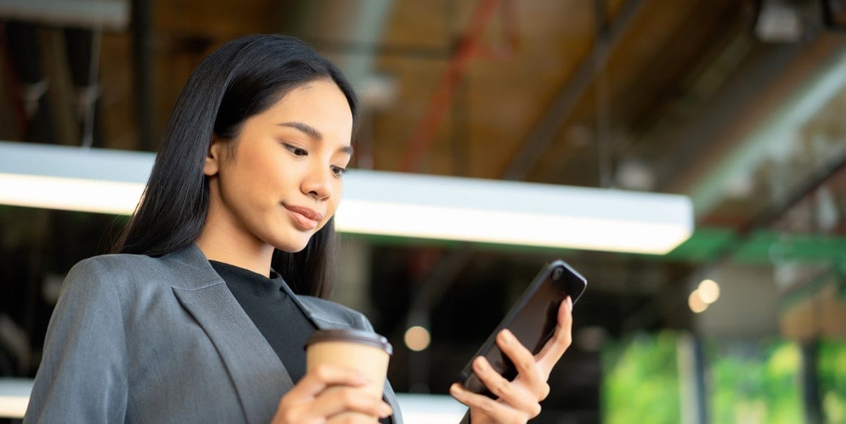 young professional woman smiling, looking at cellphone in hand, holding coffee cup